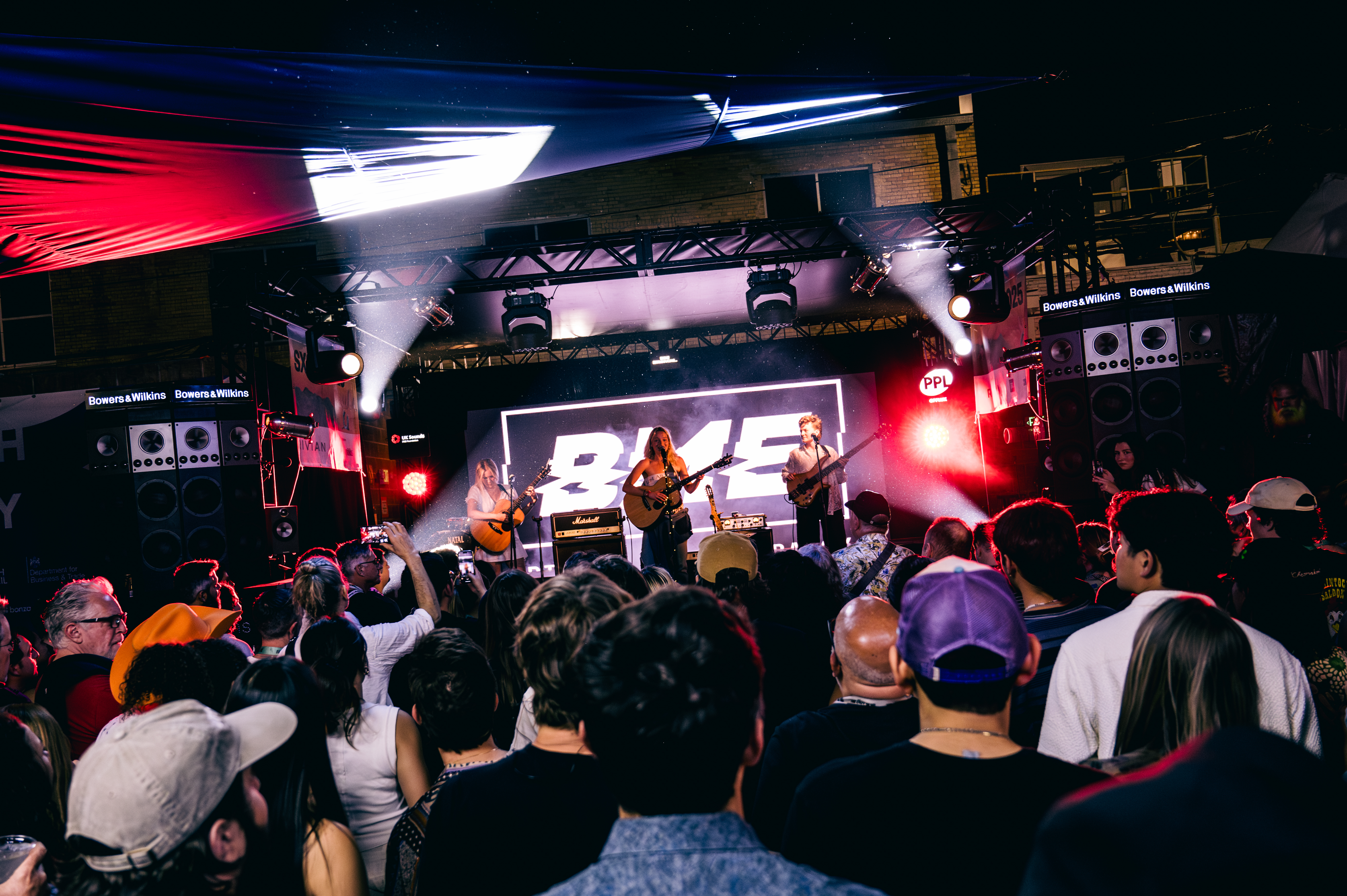 A lively crowd watches a band perform on an outdoor stage with colorful lights and a large screen displaying "BME."
